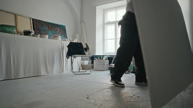 Young adult man carrying large blank canvas in art studio, placing it on floor, and sitting on chair to contemplate next painting. Ideal for creative agency promotions or art school ads