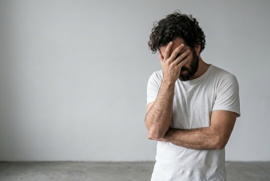 Stressed and Depressed Young Man Covering Face with Hand Standing Against a Grey Wall with Copy Space