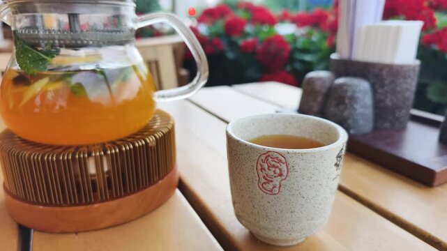 Hot tea in glass teapot creates outdoor dining atmosphere on cafe terrace. Relaxing outdoor dining setup features steaming cup on wooden table surrounded by green plants and red flowers.