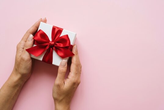 Woman Hands Holding White Gift Box with Red Ribbon Bow on Pink Background with Copy Space for Celebration
