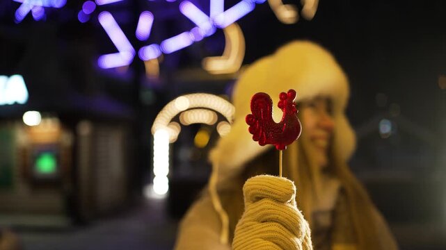 Festive woman in a fur hat and warm mittens holding a red rooster-shaped lollipop, smiling at a vibrant christmas market with bokeh lights illuminating the magical winter evening