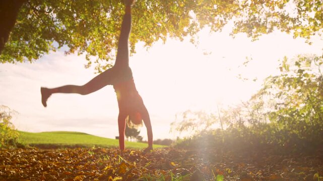 LENS FLARE, SLOW MOTION, DOF, LOW ANGLE VIEW: Cheerful woman performs a cartwheel beneath a sunlit autumn tree, surrounded by fallen leaves and warm golden light. Joy, freedom and seasonal beauty.