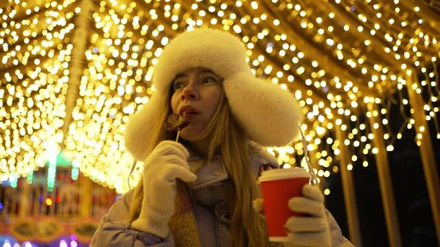 Cheerful young woman enjoying a rooster shaped lollipop and a hot drink from a red cup at a christmas market, standing under a glowing arch of festive lights on a cold winter night