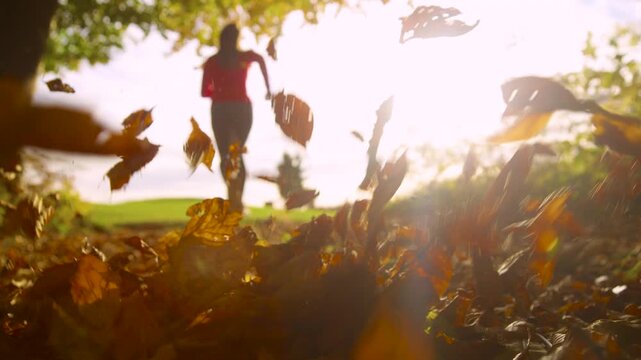 LENS FLARE, SLOW MOTION, DOF, LOW ANGLE, CLOSE UP: Young woman runs through forest path as dry autumn leaves lift into the air around her. Morning recreation on a warm sunny morning in fall season.