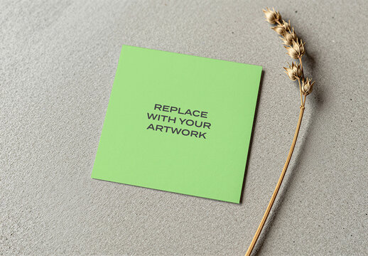 Square card mockup with replace your artwork text displayed on a textured gray surface, accompanied by dried plant stem, stationery branding presentation, print design template