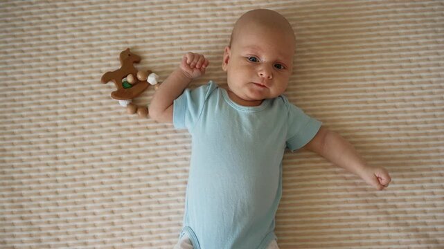 Top down view of a curious newborn baby boy lying awake on a beige blanket, wearing a blue onesie and making adorable facial expressions while actively moving his arms and legs