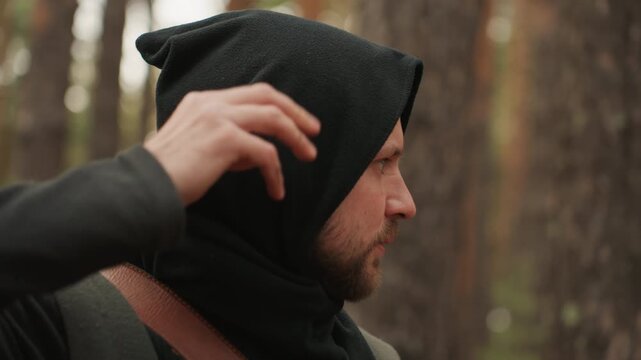 A hooded figure aiming and drawing a bow, in a forest with tall trees, with bow, arrow, from a over-the-shoulder shot from behind the hooded perspective, with black, brown accents.