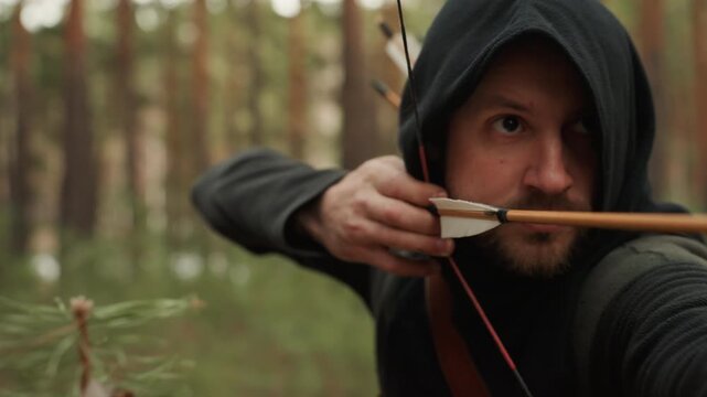 Hooded man draws back bow with arrow nocked, aiming through foreground pine branches in dense forest, holding steady shooting stance under soft daylight and natural green woodland surroundings