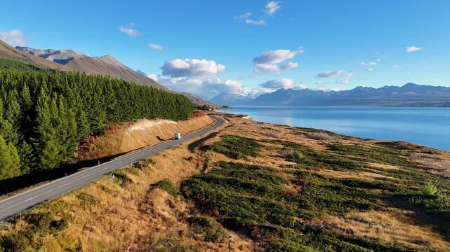 Aerial drone view of a scenic highway along Lake Pukaki with a mountain background. A camper van is driving on a beautiful lakeside road, South Island, New Zealand. 
