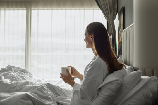 Happy Woman Enjoying Morning Coffee in Bed While Looking Out Window
