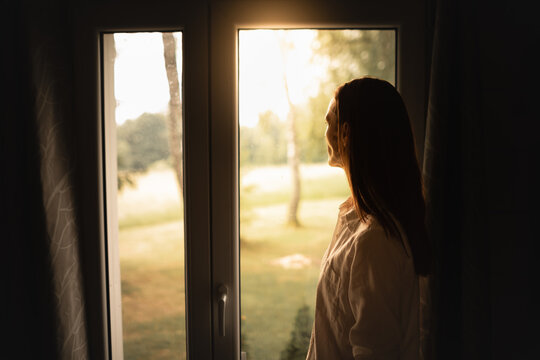 young woman looking out bedroom window watching the sunrise feeling a peace and happy 