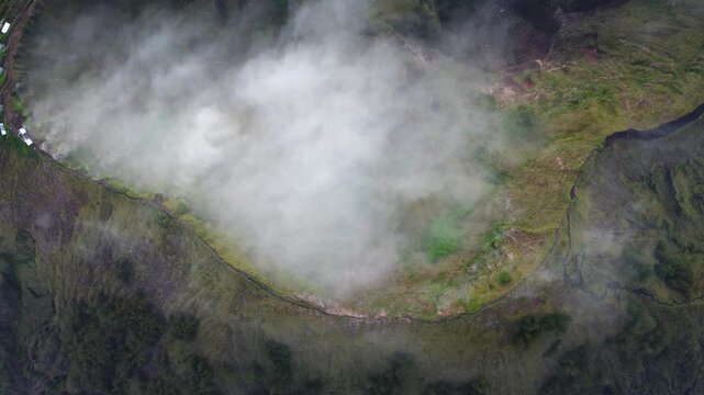 Soft mist swirls across a green crater rim in Indonesia as a drone hovers overhead in a calm top-down view.