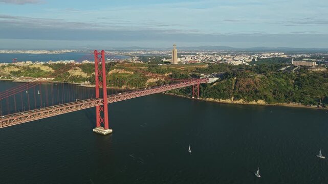 Panoramic aerial view of the 25 de Abril suspension bridge, Cristo Rei monument, Almada coastline, and sailboats on the Tagus River in Portugal.