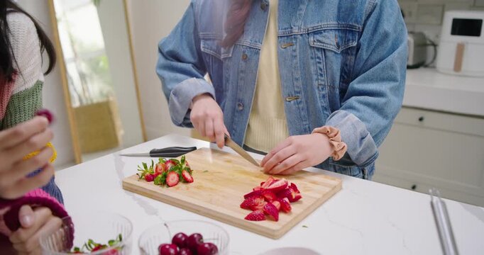 Knife being picked up is slicing strawberries into wedges on cutting board for topping dessert