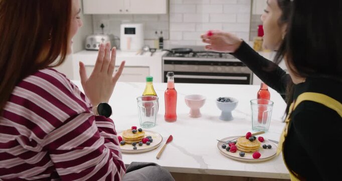 Two women friends sitting in kitchen right woman reaching berries adding to pancake taking forks