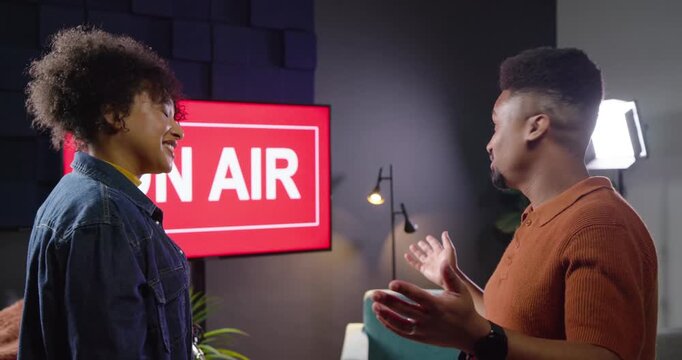 African American co-hosts responding to ON AIR sign, shaking hands, adjusting microphones, sitting