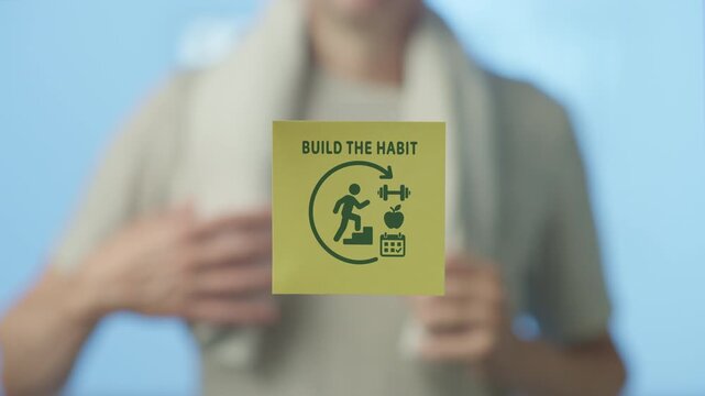 Out of focus person with a towel placing a sticky note with a build the habit message and health icons on a glass surface, symbolizing a commitment to fitness, diet, and routine