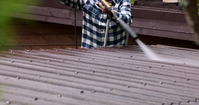 Man standing on ladder and washing barn bitumen corrugated roofing sheets with high pressure washer. Roof cleaning service