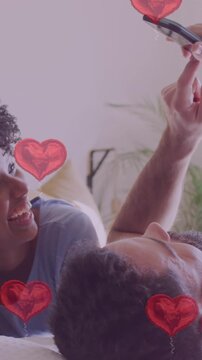 Animation of hearts over diverse couple using smartphone in bed