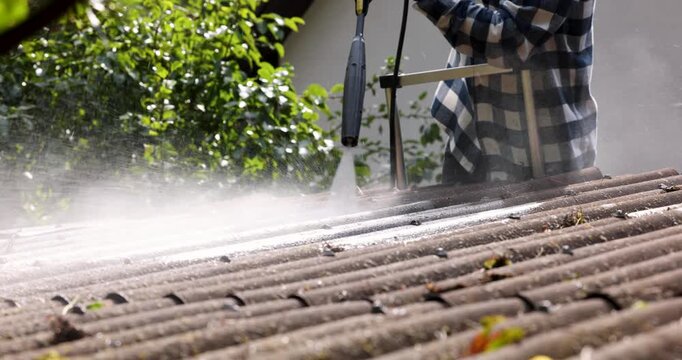 Man standing on ladder and washing dirt and moss from corrugated roofing sheets with high pressure washer. Roof cleaning service