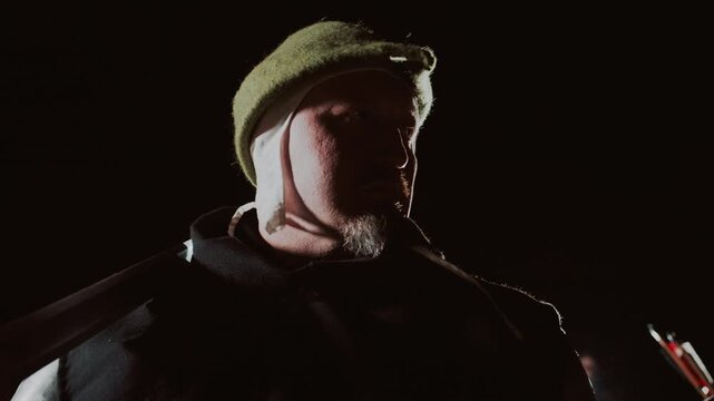 Older man in knit cap and heavy coat turns head away from camera as strong backlight creates bright rim light around cap and shoulder, leaving face mostly in shadow with moody contrast and tense