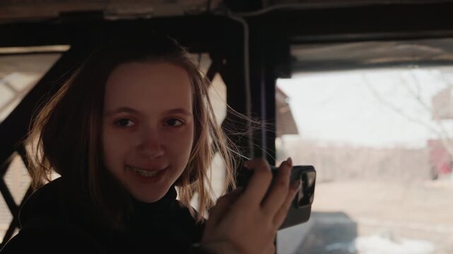 Young woman seated inside dark wooden shelter holds compact camera near chest, raises hands with gentle smile, looks slightly off camera, and shows subtle torso rotation under soft overcast daylight