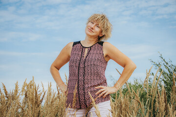 Smiling blonde woman stands with hands on hips among tall golden grasses under a soft blue sky. © Jolanta