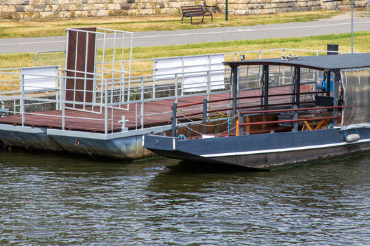 A dark-hulled converted barge moored to a floating dock alongside a stone river wall,  quay, with a transparent waterproof PVC canopy cover around. recreational houseboat as a floating storage. 