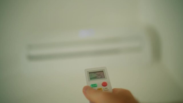 Hand holding a white air conditioner remote controller, pointing towards a blurry wall mounted air conditioning unit