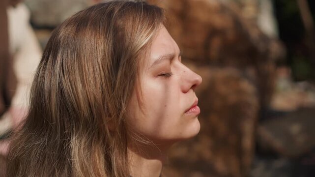 A woman poses with eyes closed in a serene outdoor setting, showcasing a side profile with trees and stone structures in natural light. This beauty and wellness lifestyle photography.