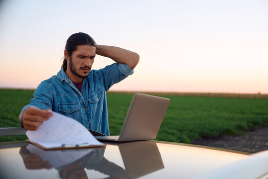 Distraught farm owner analyzing reports while using laptop in agricultural field.