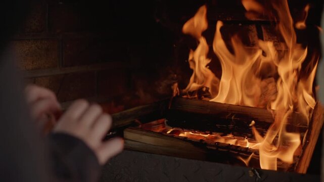 Hands held near active indoor wood fireplace as tall orange-yellow flames rise over stacked charred logs, glowing embers and soft sparks creating warm light, cozy comfort, visible heat, gentle