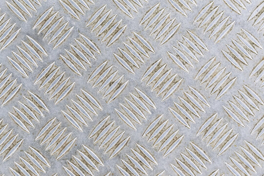Silver diamond plate metal texture. Raised diagonal tread pattern across industrial surface. Durable aluminum flooring material detail. Geometric non slip design. Metallic industrial background.
