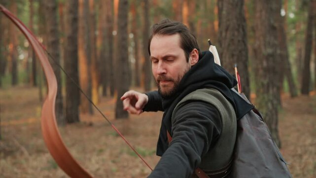 A man is seen aiming and drawing back the bowstring in a forest setting, showcasing a focused and determined expression. This scene is part of an outdoor archery activity, reflecting.