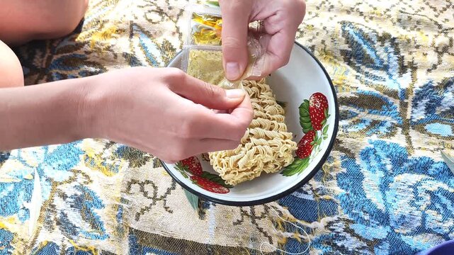  Person pouring instant ramen noodles into white bowl with fresh strawberries on colorful fabric, outdoor picnic setting.