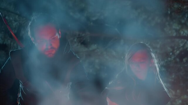 People bend toward campfire glow at night, heads lowered and gazes fixed on embers while smoke drifts upward, creating warm firelight against cool shadows in wild setting, with campfire, smoke, night