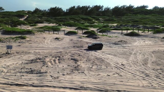 Off road vehicle crosses golden beach dunes at sunset, moving from left to right as the camera follows its path. Miramar Beach on the Gulf of Mexico provides a tropical setting with green coastal vege