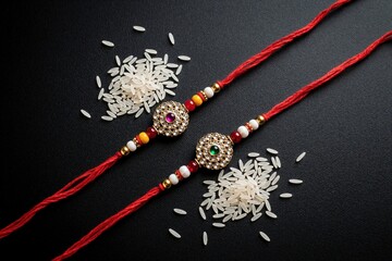 Fototapeta premium Two decorative rakhi threads with beads, jewels lie on black background. White rice grains scattered around festive bracelets. Raksha Bandhan celebration items for Indian sibling love, traditional