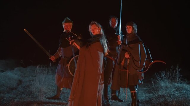 Four armored warriors move toward camera through dark mist, central cloaked leader lifting sword upright while companions hold spears and round shield, creating dramatic battle tension