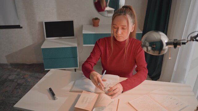 Young woman in red top is seated at desk writing. Woman untangles white corded headphones while handling various. Setting features light colored walls patterned carpet blue striped