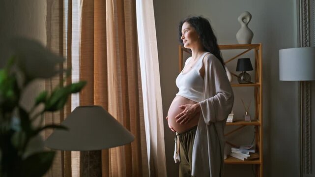 Expectant woman cradling belly window home. Mother gazing outside cozy interior.