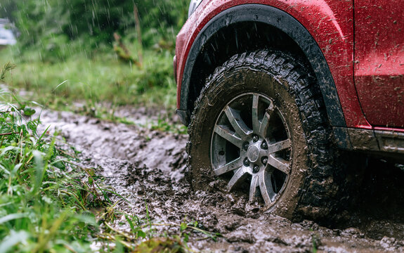 Red car wheel stuck in deep wet mud on dirt road during heavy rain. Dirty automobile tire sink in rural pathway landscape with green grass. Vehicle struggle in slippery clay terrain offroad travel