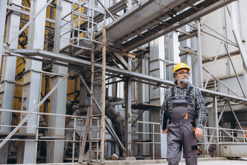 Fototapeta premium Male worker wearing hard hat and uniform, carrying a tablet, supervising operations at a large grain processing plant