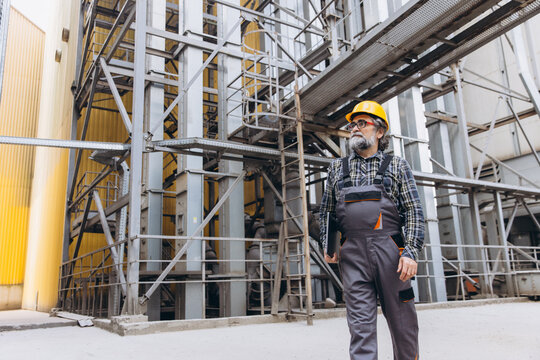 Male worker with beard and safety gear walking on an industrial site with grain silos and metal structures