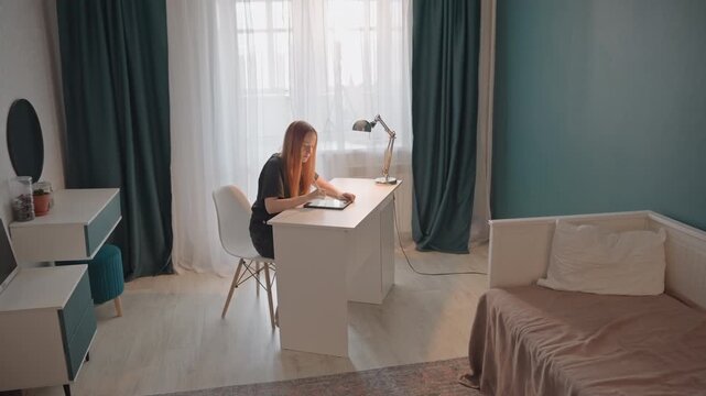 Young woman with long red hair works at white desk in well lit. Room features teal walls dark green curtains. Desk setup includes silver adjustable lamp small round mirror