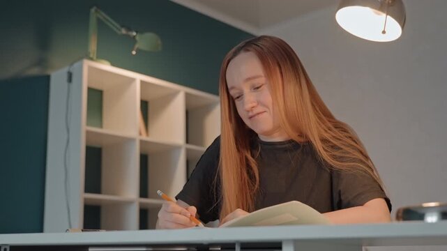 Young woman with long hair is seated at desk writing. Woman wears dark top and uses orange pencil while another pen. Background features white shelves filled showing woman