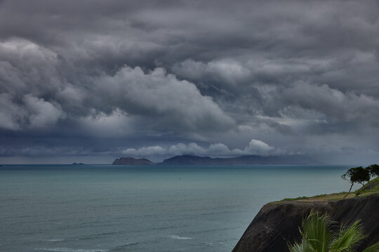 storm clouds roll over San Lorenzo Island as seen from the cliffs of Miraflores, Lima, casting a moody, atmospheric veil over the Pacific coastline. The dense cloud formations and shifting light hint 