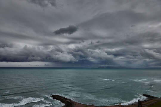 storm clouds roll over San Lorenzo Island as seen from the cliffs of Miraflores, Lima, casting a moody, atmospheric veil over the Pacific coastline. The dense cloud formations and shifting light hint 