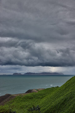 storm clouds roll over San Lorenzo Island as seen from the cliffs of Miraflores, Lima, casting a moody, atmospheric veil over the Pacific coastline. The dense cloud formations and shifting light hint 