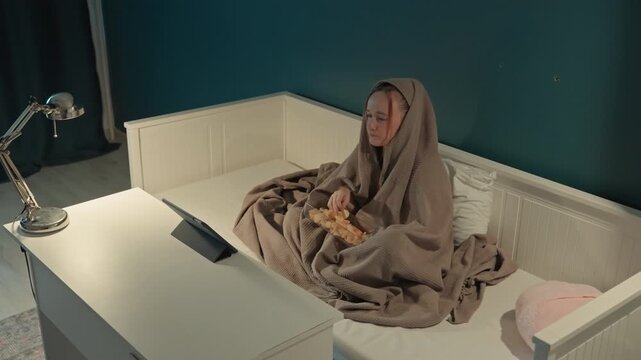 Young woman sits on white daybed covered with brown blanket eating. Silver desk lamp and pink pillow are. Scene remains consistent throughout showing minimal movement